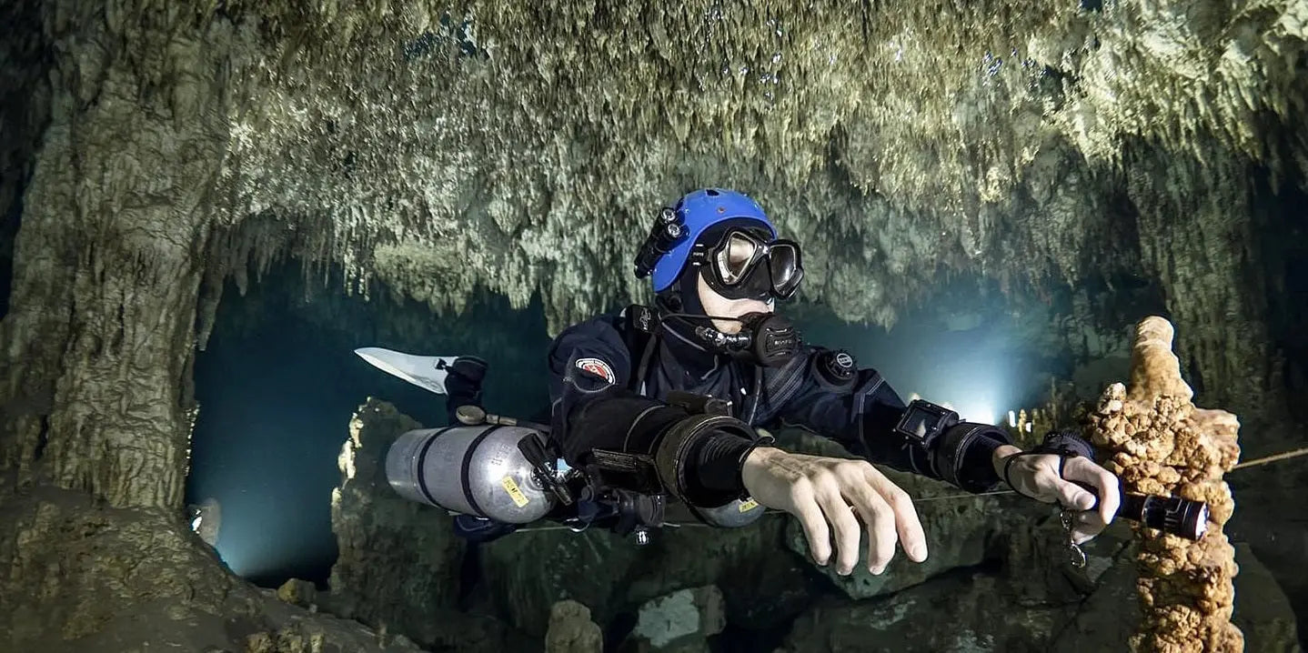 Cave diver Marcelin Nebenhaus wearing a blue helmet and black gear, exploring an underwater cave with a flashlight, surrounded by stalactites and rock formations.