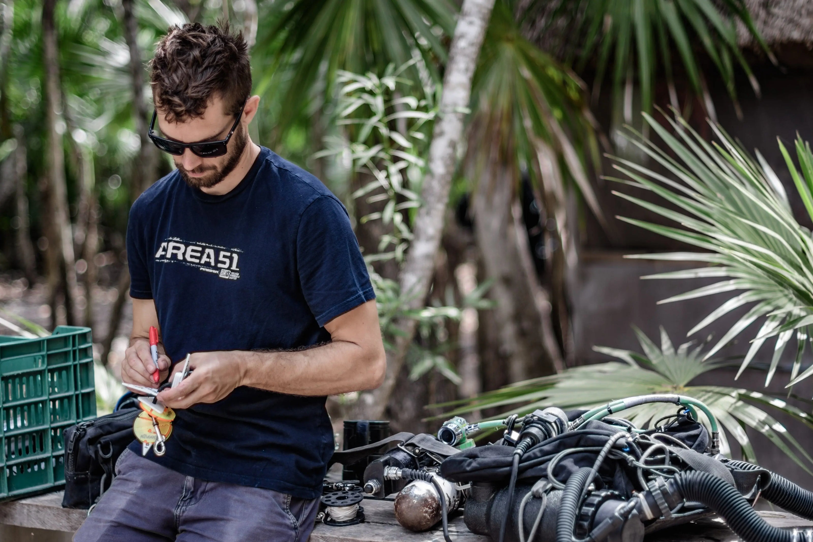 RAID Certified Instructor Skanda Coffield-Feith wearing a dark blue t-shirt and sunglasses, working on scuba equipment at a table in a tropical setting with palm trees in the background.