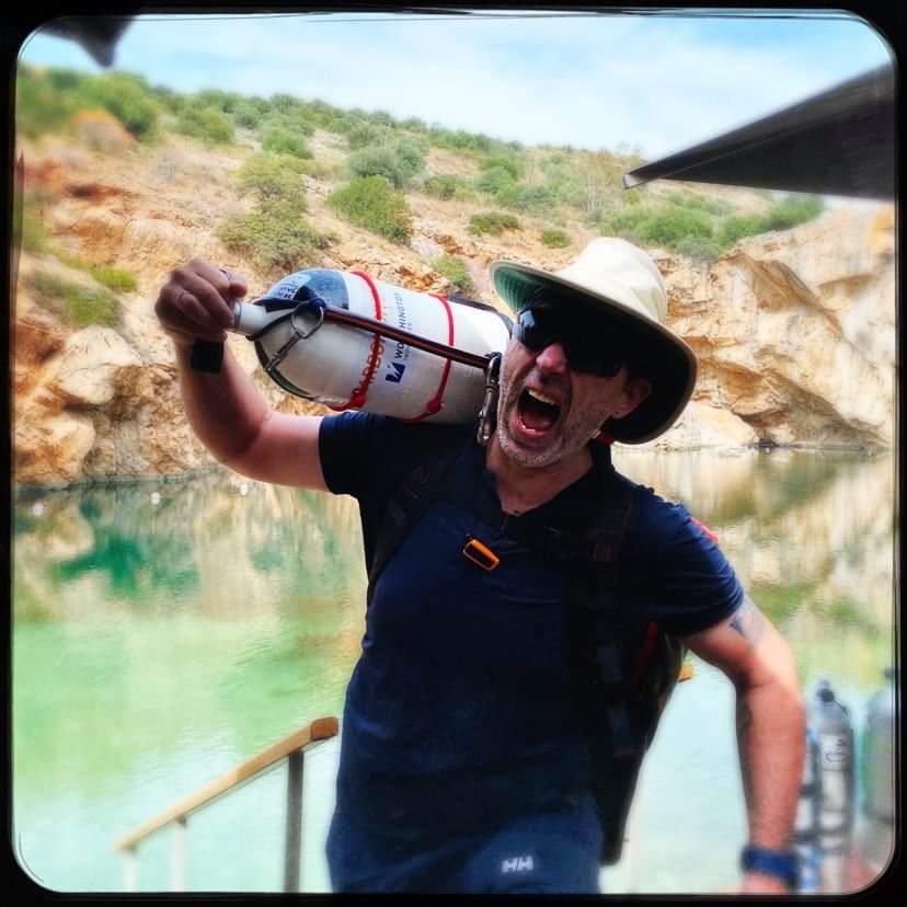 Diver Ioannis Dalmiras holding a scuba tank on his shoulder, wearing a wide-brimmed hat and sunglasses, with a rocky and green-water background.